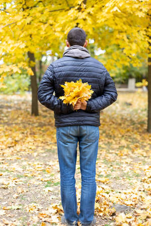 A man in a black jacket holds a bouquet of yellow autumn maple leaves behind his back. rear view. autumn moodの写真素材