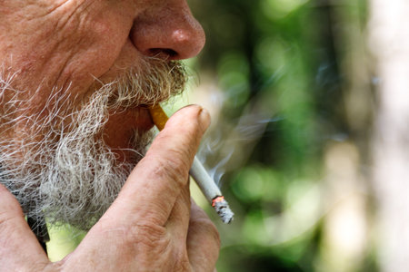 Old man with wrinkles and beard smoking a cigarette outdoor. Closeup hands holding a cigarette with smokeの写真素材