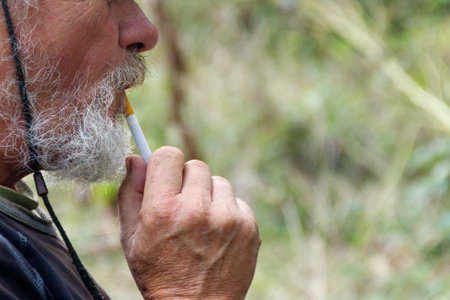 Old man with wrinkles and beard smoking a cigarette outdoor. Closeup hands holding a cigarette with smokeの写真素材