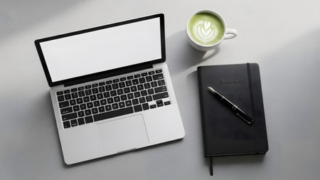 A laptop, black notebook with a pen, and a cup of matcha latte sit on a gray desk surface.