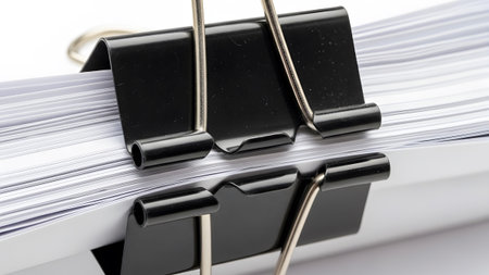 showing close up of a black binder clip holding a stack of white paper sheets on a white...