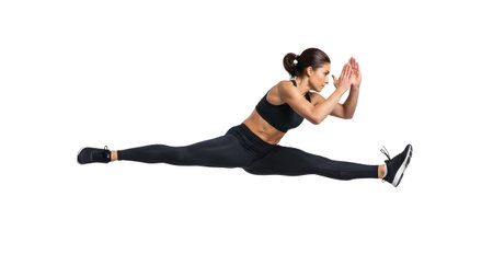 A woman in a black sports bra and leggings performs a wide split jump with hands clasped together.