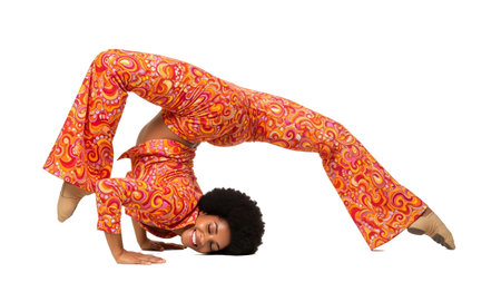 A woman in a vibrant orange 1970s patterned outfit performs a challenging yoga pose on a white...