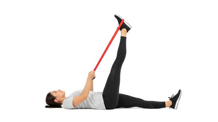 A woman lies on her back, pulling a red resistance band attached to her foot for a leg stretch.