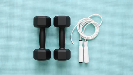 Two black dumbbells and a white jump rope are arranged on a light blue textured yoga mat base.