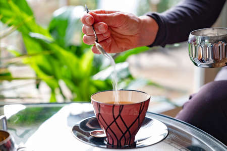 Women's hand does sugar in the pink retro tea cup, behind her a window through which the sun shines.