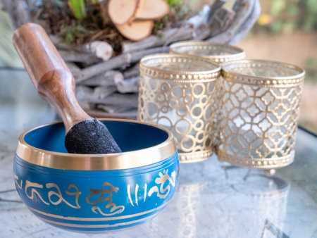 Haulerwijk - march 07 2020: Haulerwijk, The Netherlands. blue and golden indian singing bowl made of seven metals with a wooden striker on a glass table, plants in backgroundのeditorial素材