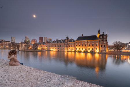 The Hague - February 17 2019: The Hague, The Neherlands. Binnenhof castle, Dutch Parliamen, with the hofvijver, a young woman with brown hair is sitting on the quay , a blue sky with the moon, The Hague, Den Haag, The Netherlandsのeditorial素材