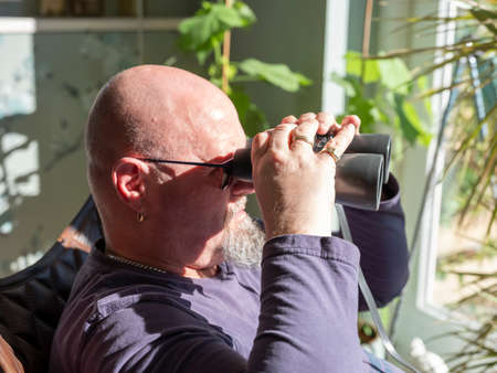 Elderly man sittiing, looking through a glass door with binoculars as he watches something in the...