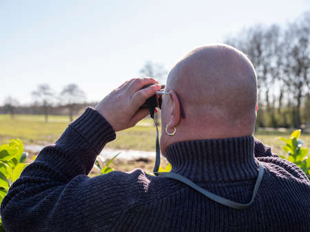 Elderly bald man standing outside, looking with the binoculars over the meadows in search of wildlifeの写真素材