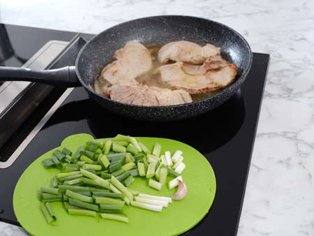 frying pan on an induction plate in which pork chops are baking, green cutting board with spring onions in foreground.の写真素材