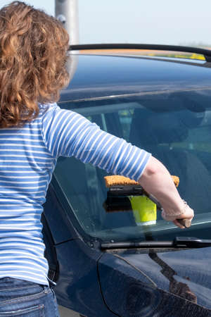 Woman cleans front glass of the car with a sponge and water. Yelllow paper cup in the car with text....