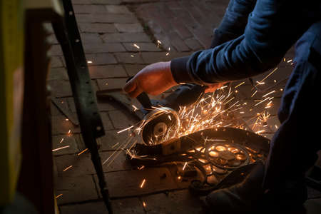 Side view of a mans hands working on a metal part of a garden bench, using an electric grinder while sparks are flying around in the industrial workshopの写真素材