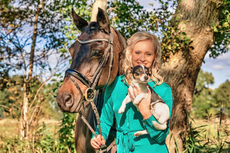 Beautiful portrait of a blond smiling girl with her horse and dog in the forest. Wearing a green dress. Selective focus.の写真素材