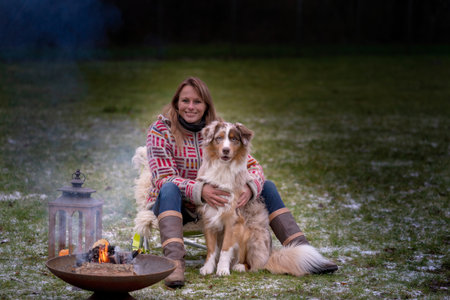 An Australian Shepherd sits by the campfire with his owner in the afternoon. Candles burn in a lantern. Woman and dog cuddle and enjoy camping life, in winterの写真素材