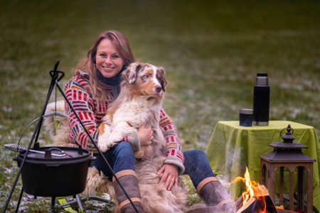 Tricolor Australian Shepherd sits next to a young woman by the campfire. In winter, snow on the grass. The fire is burning under the cauldron.の写真素材