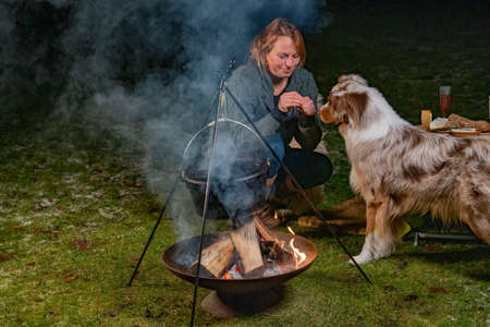 Young smiling blond woman gives biscuits to her tricolor Australian Shepherd dog. In winter, next to the smoking kettle over the campfire, in the dark outside in the park. holiday, camping, weekend, animal themes.の写真素材