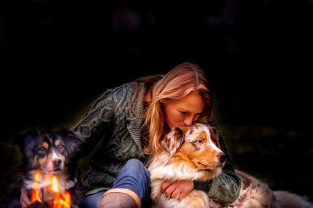 Young woman is sitting outside in the woods with her two Australian Shepherd dogs. Snow on the grass, at night by the campfire. She kisses the dog lovingly.の写真素材