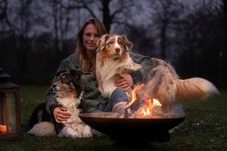 Young woman is sitting outside in the woods with her two Australian Shepherd dogs. Snow on the grass, at night by the campfire. She embraces the dogs lovingly.の写真素材