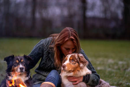 Young woman is sitting outside in the woods with her two Australian Shepherd dogs. Snow on the grass, twilight by the campfire. Cuddles and companion animalsの写真素材