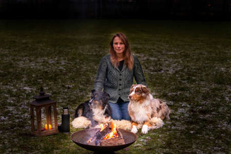 Young woman is sitting outside in the woods with her two Australian Shepherd dogs. Snow on the grass, at night by the campfire. She embraces the dogs lovingly.の写真素材