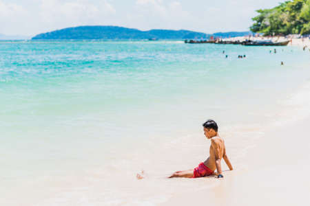 Vacation in Thailand: young man in swimming trunks sits on white sand in surf line of turquoise sea against beautiful seascape of Railay Phra Nang Beach, Krabi. Concept of travel to tropical paradise.の写真素材