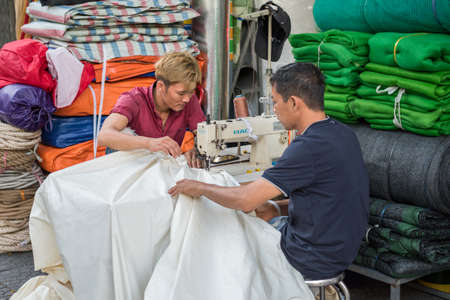 Ho Chi Minh City, Vietnam - February 11, 2019: Vietnamese men stitch a fabric with a sewing machine in the street near their small shop. Saigon is abundant with such small street businesses.のeditorial素材