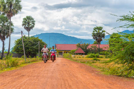 Kampot, Cambodia - December 14, 2016: local men holding fishing rods drive motorbikes along a rural dirt road.のeditorial素材