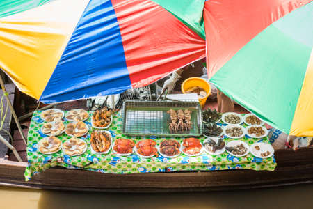 Amphawa, Samut Songkhram / Thailand - June 15, 2019: a table with grilled seafood below colorful umbrellas in a boat at Amphawa Floating Market.のeditorial素材