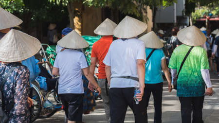Hoi An, Vietnam - October 23, 2018: group of Asian tourists in Vietnamese conical hats walk in the street, view from behind. The number of Chinese travelers has significantly grown recently in Hoi An.のeditorial素材
