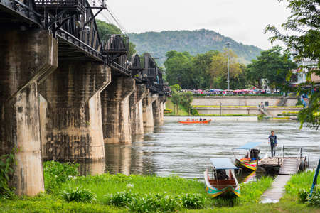 Kanchanaburi, Thailand - August 18, 2018: The bridge on the River Kwai (Khwae Yai), crowds of tourists on the distant embankment, boats on the river and a small pier adjacent to a floodplain lawn.のeditorial素材