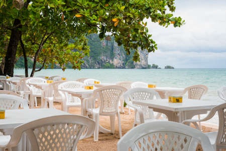 Ao Nang, Thailand - July 2, 2018: empty tables of an open-air cafe on the Ao Nang Beach against the cloudy sky and grayish sea. Summer is the low season in Krabi province so tourist business idles.のeditorial素材