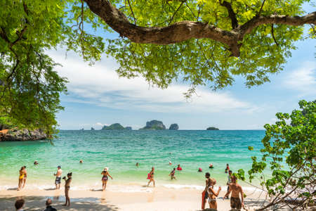 Railay, Thailand - July 3, 2018: tourists swim in the turquoise sea water of Phra Nang (Phranang) Beach with a massive tree branch above the scene & group of islands (Ko Poda & others) on the horizon.のeditorial素材