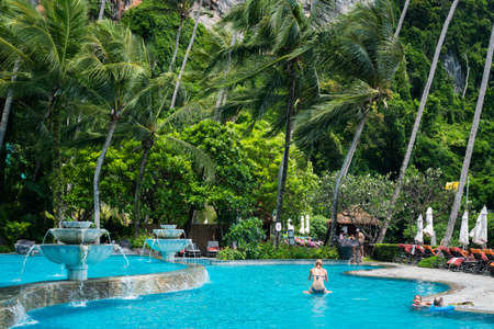 Ao Nang, Thailand - July 2, 2018: a blue pool with tourists in it below coconut trees that sway in the wind at Pai Plong Beach, Centara Grand Beach Resort & Villas Krabi.のeditorial素材
