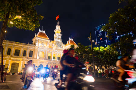 Ho Chi Minh City, Vietnam - May 1, 2018: People drive motorbikes by illuminated Saigon City Hall, Vincom Center towers and colorful tropical plants against night sky.のeditorial素材