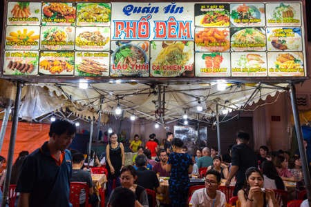 Ho Chi Minh, Vietnam - April 28, 2018: open air night street restaurant at Ben Thanh Market. Signboard shows the menu with pictures & dishes names in Vietnamese and English. People sit at red tables.のeditorial素材