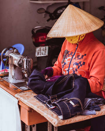 Nha Trang, Vietnam - May 5, 2018: a tailor in a rice hat and a mask works with jeans at the table with an old sewing machine at a small street workshop.のeditorial素材