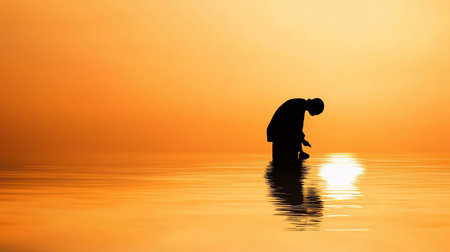 A lone, silhouetted figure stands in shallow water, head bowed, against a vibrant orange sunset. The sun's reflection creates shimmering patterns on the water's surface.の素材