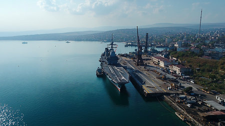 Aerial view of an aircraft carrier docked at a port, with a cityscape in the background under a partly cloudy sky. The scene is bathed in natural light.の素材