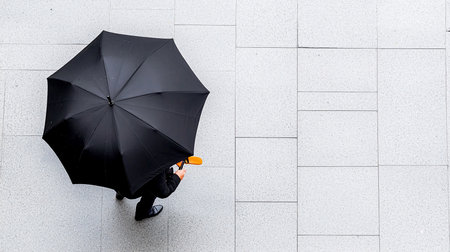 An aerial shot captures a person in business attire holding a black umbrella on a textured white pavement, creating a minimalist and modern composition.の素材