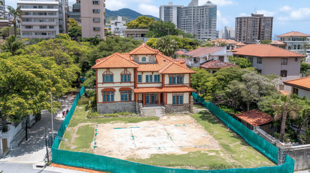 A stately colonial-style house with an orange tile roof and blue-framed windows stands in an urban landscape, surrounded by a green fence and city buildings.の素材
