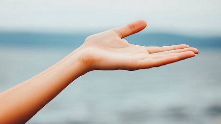 A woman's open palm extends towards the viewer, set against a blurred backdrop of soft blue hues, suggesting water or sky. The gesture implies offering, receiving, or holding space.の素材