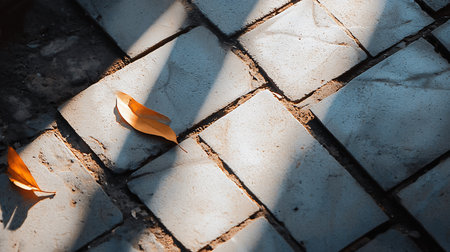 Two golden leaves rest on a gray brick path, bathed in sunlight and shadow. The herringbone pattern of the bricks adds depth to the scene, evoking a sense of tranquility.の素材