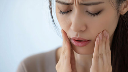 A close-up of a distressed woman with closed eyes and a furrowed brow, holding her jaw with both hands due to intense dental pain.の素材