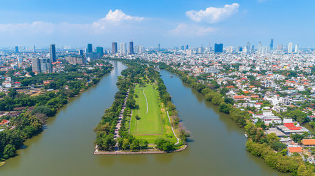 A high-angle shot of a dense city with modern skyscrapers, bisected by a wide river containing a long, lush green island with trees and manicured lawns.の素材
