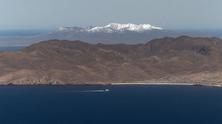 A vast mountain range with snow-covered peaks, brown rocky terrain, and a serene blue sea. The mountains have a rough texture.の素材