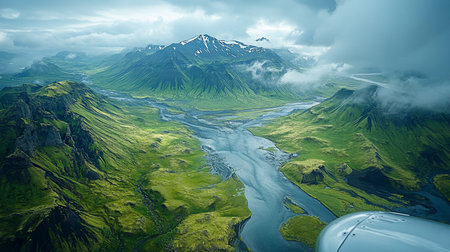 Vast aerial landscape featuring vibrant green, moss-covered mountains and valleys. A wide, winding glacial river flows through, with distant snow-capped peaks under a cloudy sky.の素材