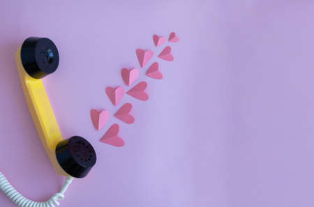 The handset of a yellow retro phone on a pink background. Pink hearts come out of the handset of the...