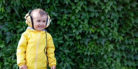 A positive little boy of 3 years old in a yellow jacket and yellow retro earphones listens to music. The boy stands against the background of a wall of plants. Banner with space for textの写真素材