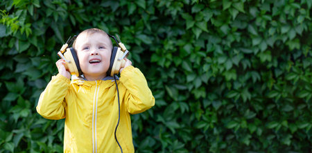 A positive little boy of 3 years old in a yellow jacket and yellow retro earphones listens to music. The boy stands against the background of a wall of plants. Banner with space for textの写真素材